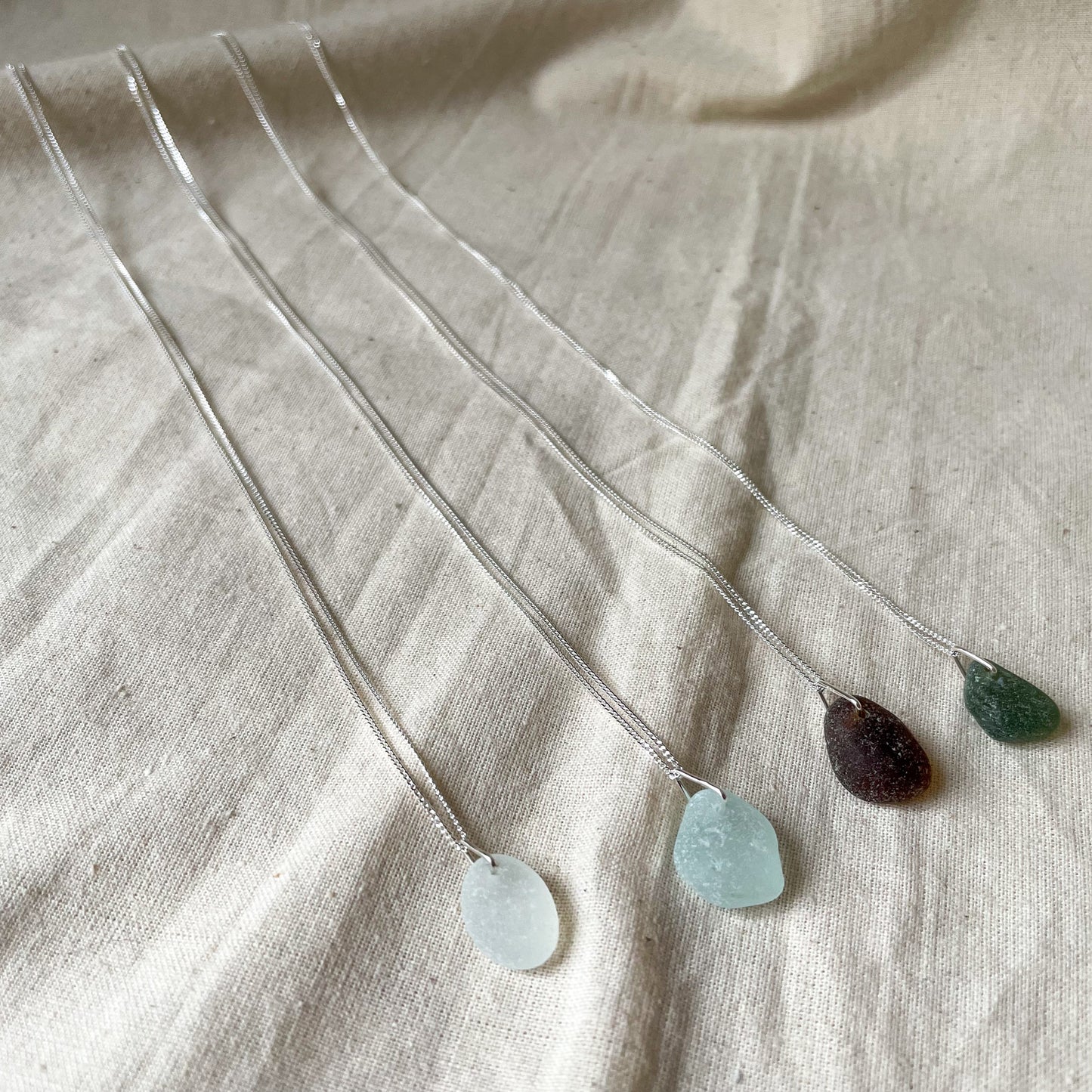 Four sea glass necklaces on fine sterling silver chains agaisnt a beige background. White sea glass necklace (left), light blue sea glass necklace (middle), brown sea glass necklace (middle) and green sea glass necklace (right)