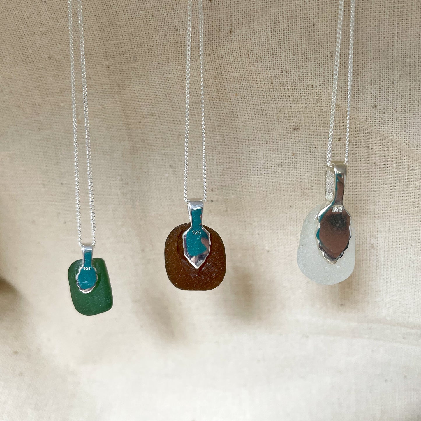 Three sea glass necklaces hanging in front of a beige background. Green sea glass necklace on the left, brown sea glass necklace in the middle and white sea glass necklace on the right.