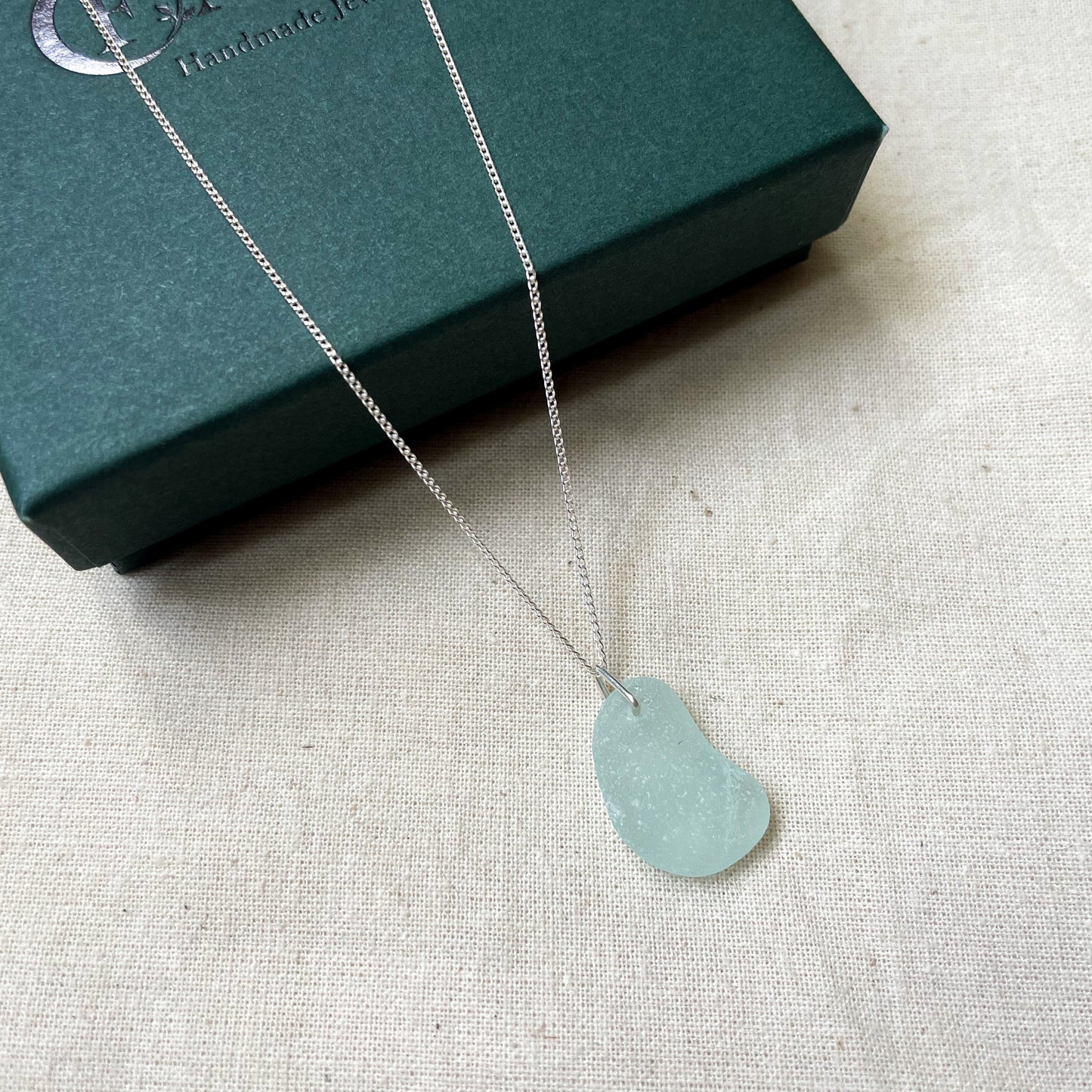 Blue sea glass necklace on fine silver chain hanging over a dark green gift box