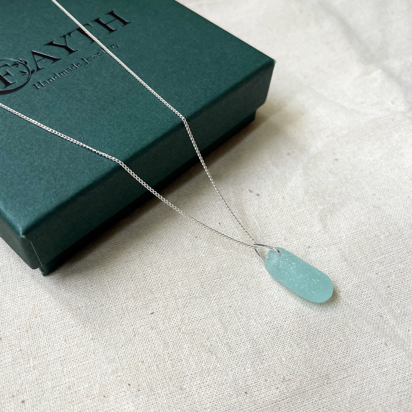 Blue sea glass necklace on fine silver chain hanging over a dark green gift box