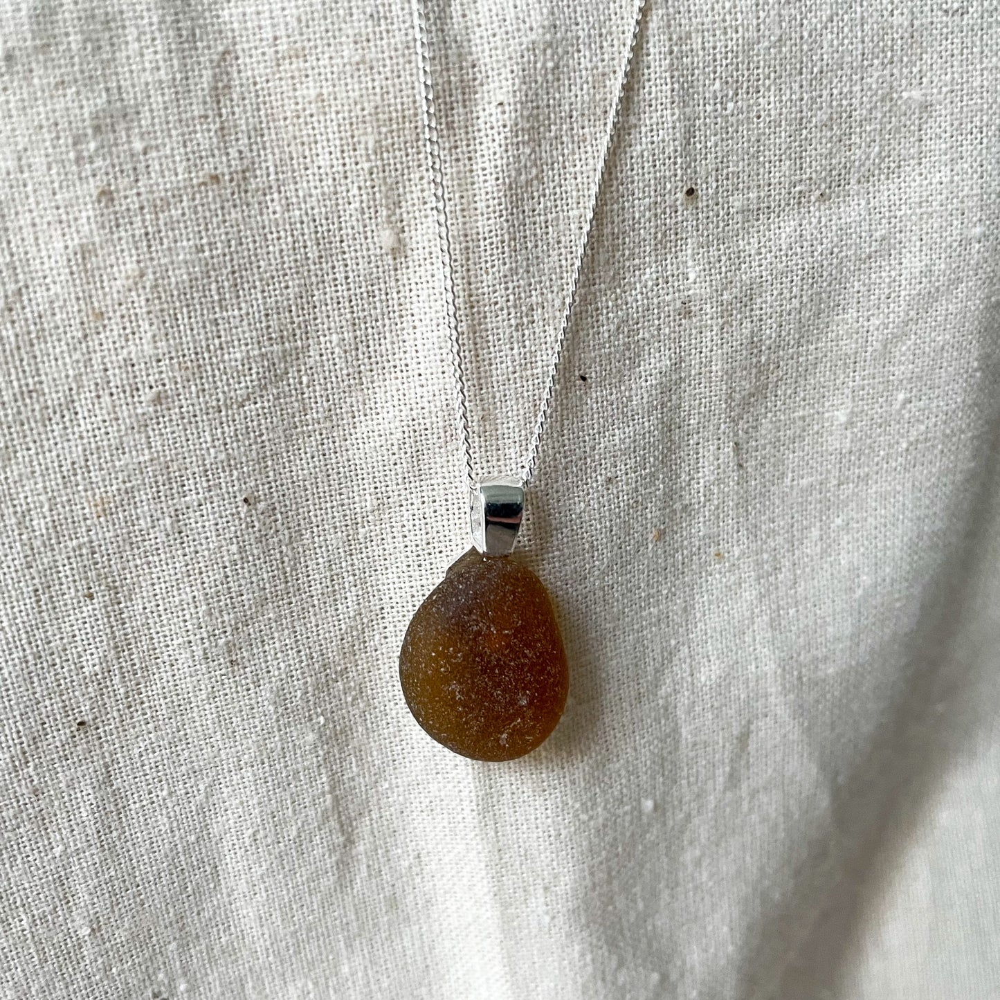 Light Brown sea glass and sterling silver necklace hanging against a beige background