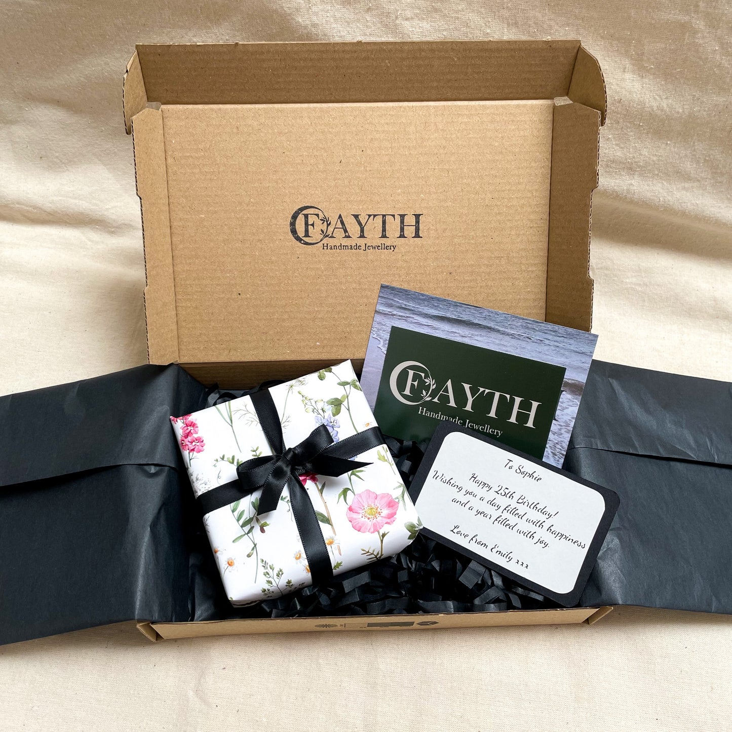 Gift box with black ribbon inside a brown cardboard box with black tissue paper, business cards and note card
