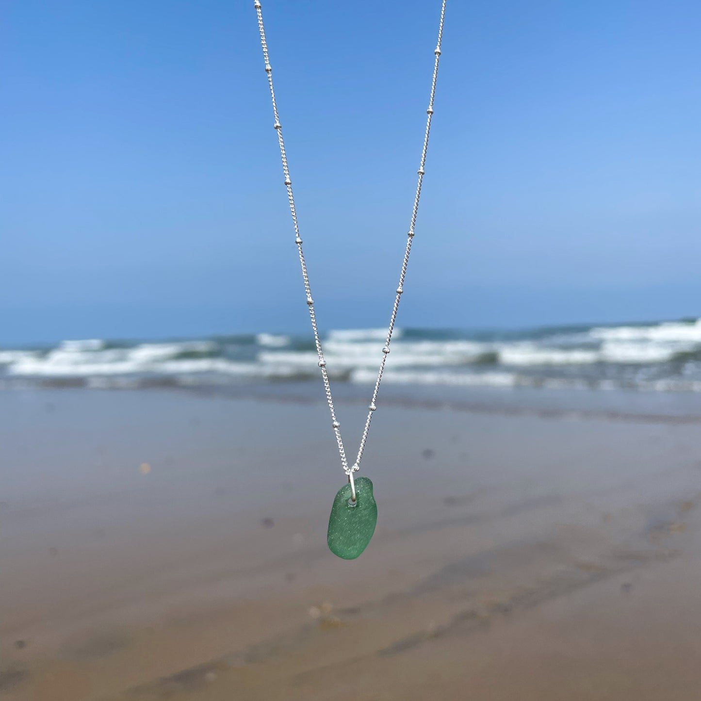 green sea glass and sterling silver necklace hanging in front of a blurred background of the sea.