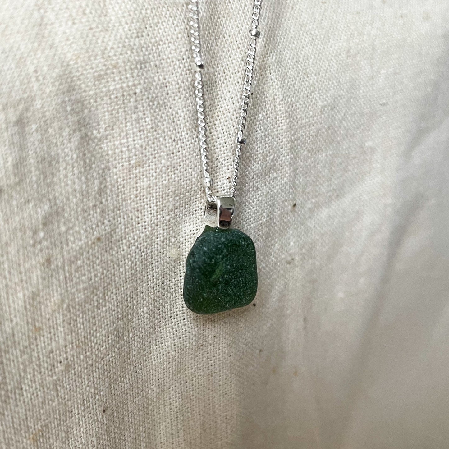 Green sea glass necklace on a sterling silver satellite chain hanging against a beige background