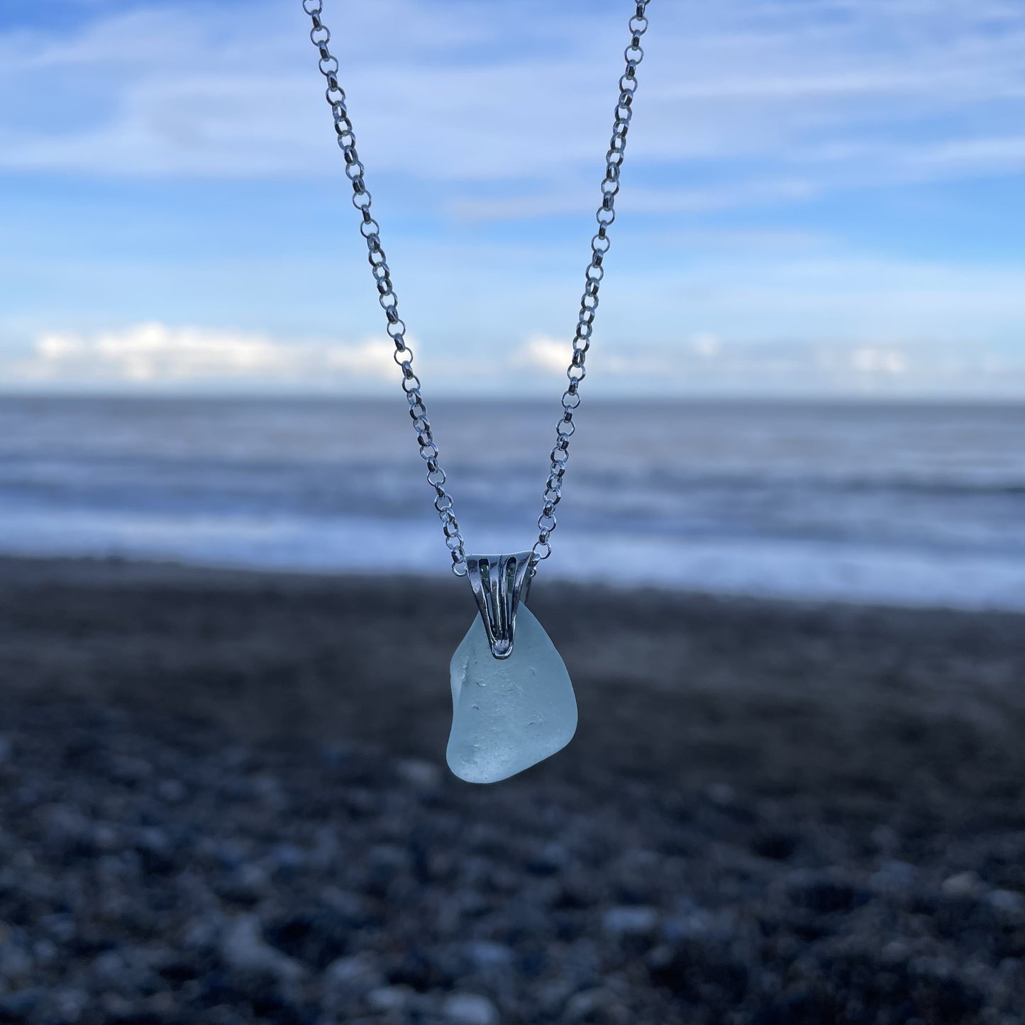 light blue sea glass pendant on a sterling silver belcher chain hanging in front of a blurred background of the sea.