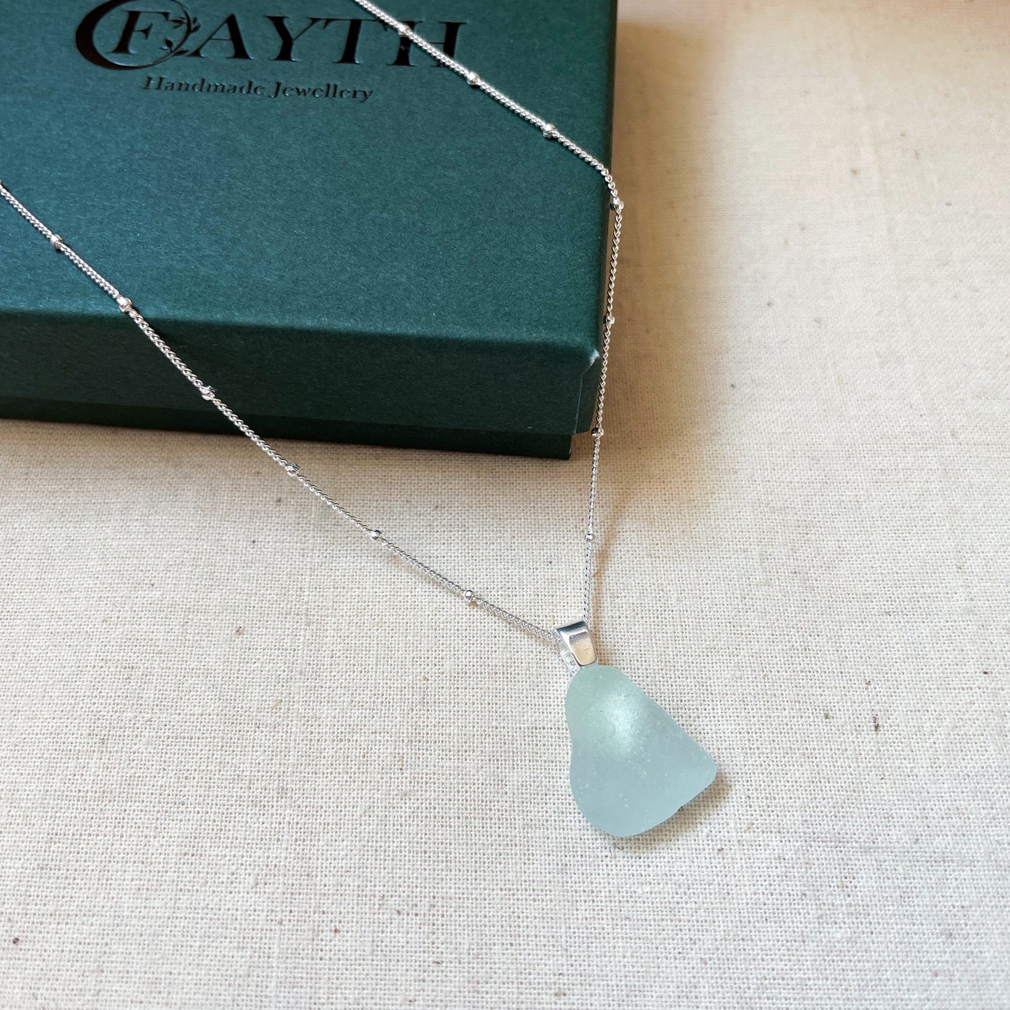 Light blue sea glass necklace on silver satellite chain hanging over a dark green gift box