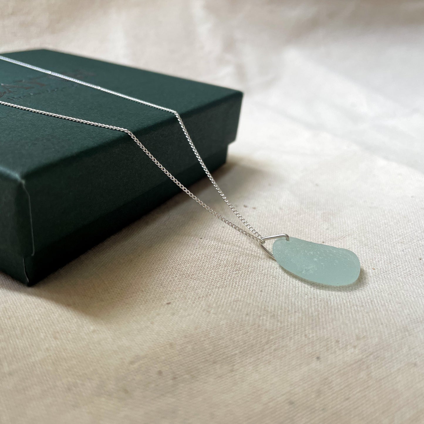 Light blue sea glass necklace on fine silver chain hanging over a dark green gift box