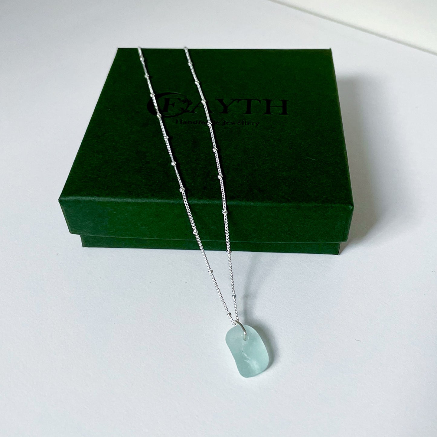 light blue sea glass necklace on silver satellite chain hanging over dark green gift box