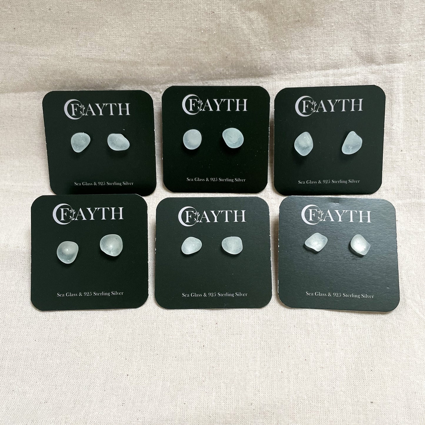 six pairs of very light blue sea glass studs on branded dark green square display cards, in two rows of three.