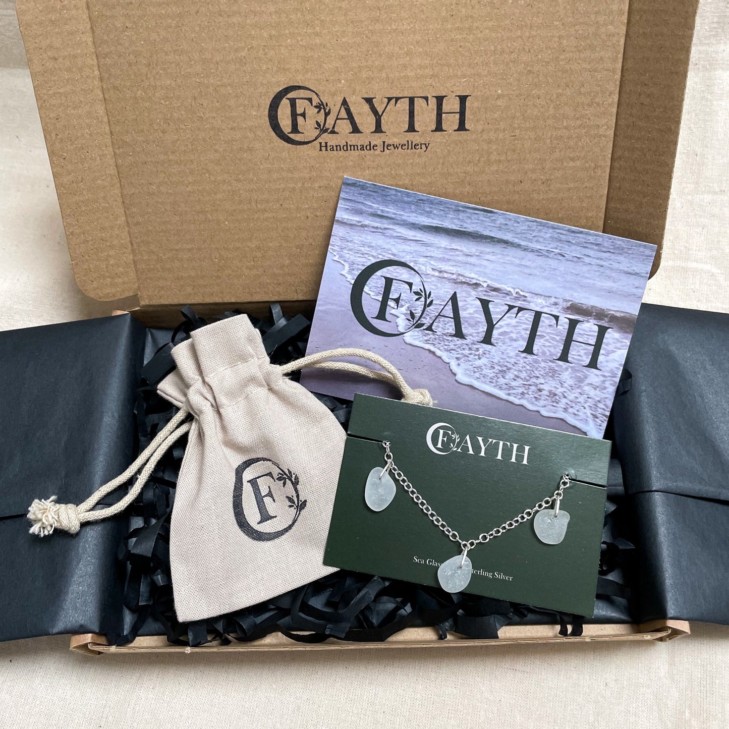 White sea glass bracelet on a branded dark green display card is inside a brown cardboard box with black tissue paper, a branded beige cotton tie gift bag and a business card.