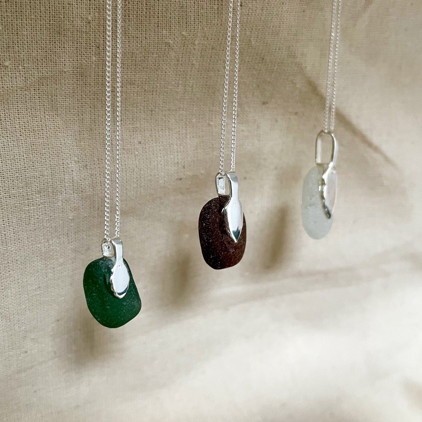 Side/back of three sea glass necklaces hanging in front of a beige background. Green sea glass necklace on the left, brown sea glass necklace in the middle and white sea glass necklace on the right.