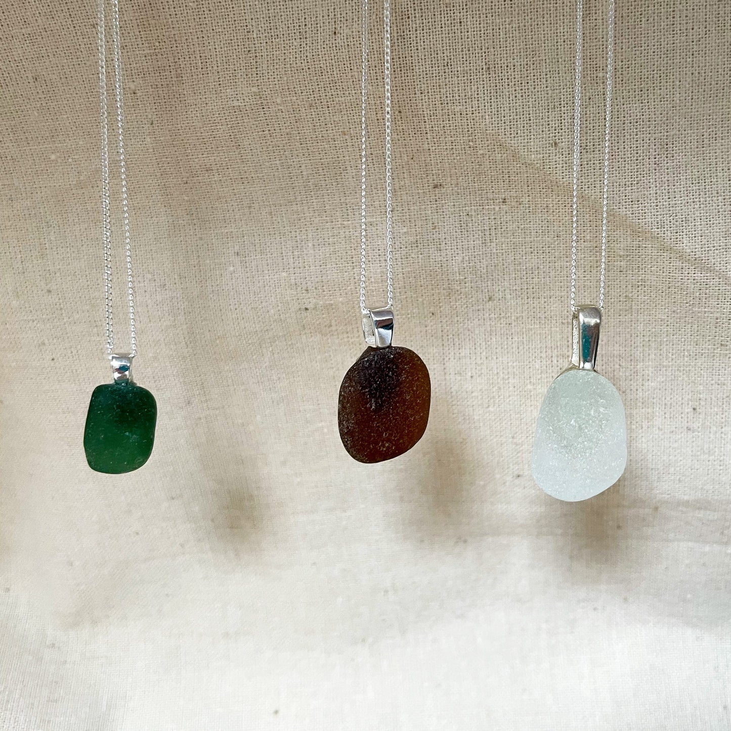 Three sea glass necklaces hanging in front of a beige background. Green sea glass necklace on the left, brown sea glass necklace in the middle and white sea glass necklace on the right