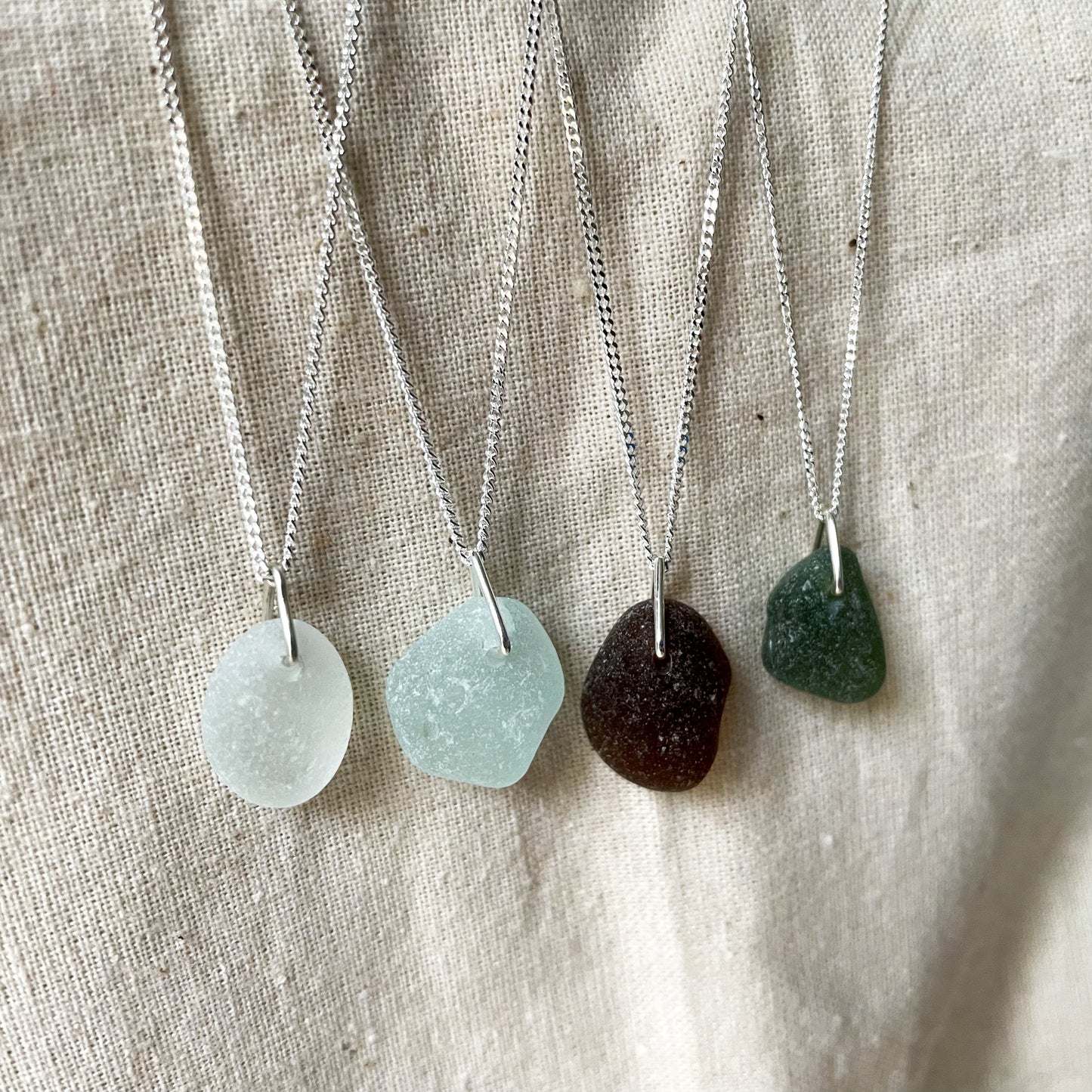 Four sea glass necklaces on fine sterling silver chains hanging agaisnt a beige background. White sea glass necklace (left), light blue sea glass necklace (middle), brown sea glass necklace (middle) and green sea glass necklace (right)