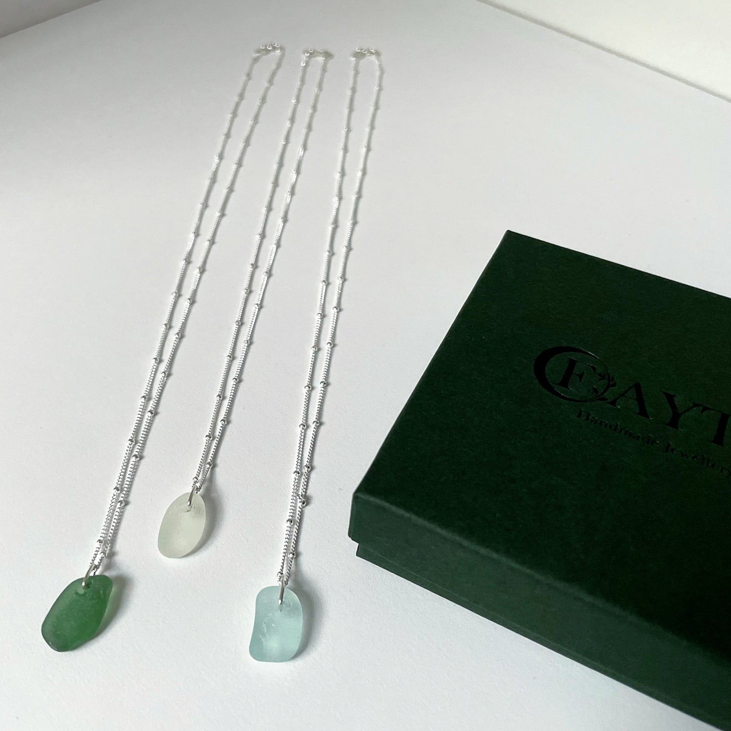 Three sea glass necklaces on silver satellite chains with dark green gift box to the right side. Green sea glass necklace (left), white sea glass necklace (middle) and light blue sea glass necklace (right).