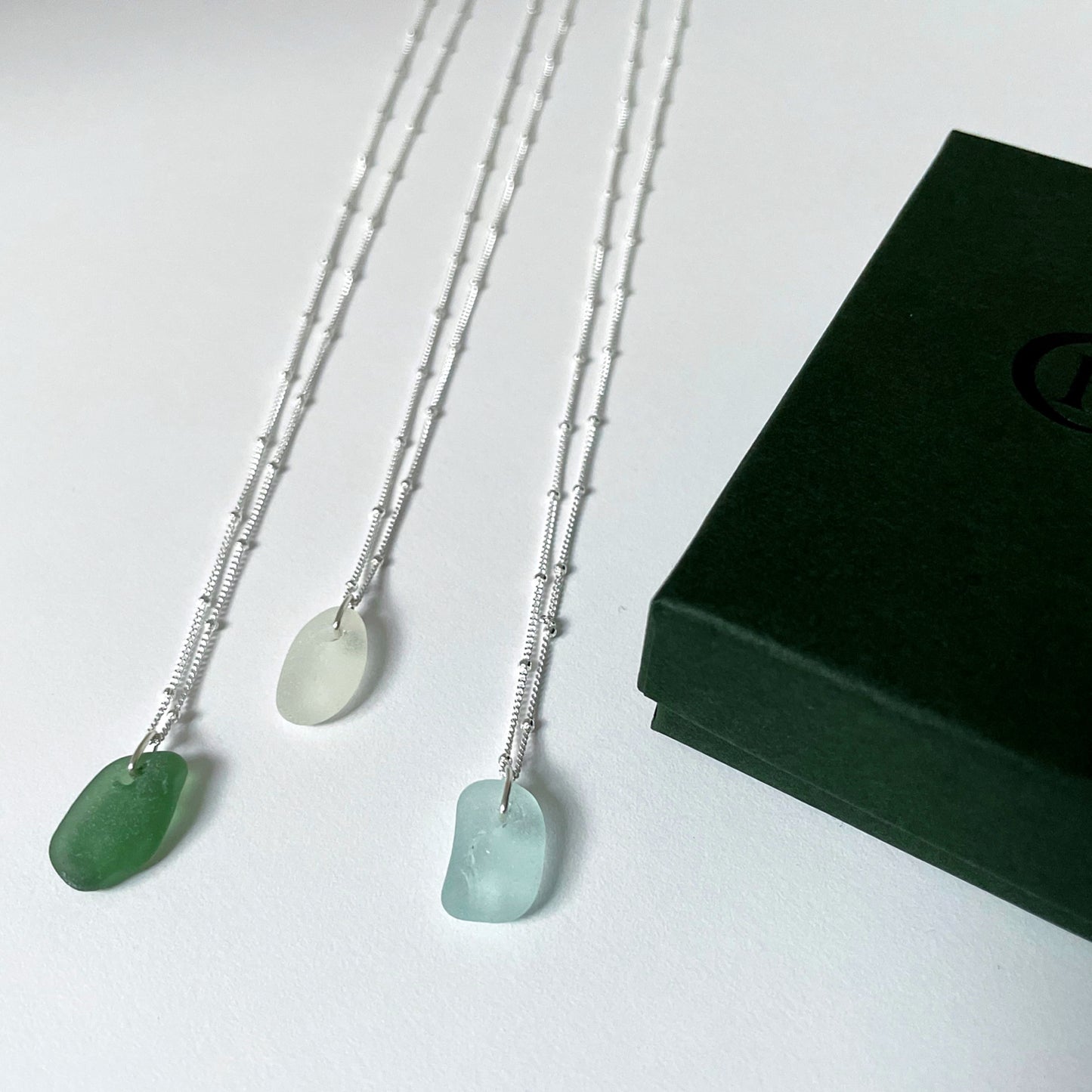 Three sea glass necklaces on silver satellite chains on a white background with dark green gift box to the right side. Green sea glass necklace (left), white sea glass necklace (middle) and light blue sea glass necklace (right).
