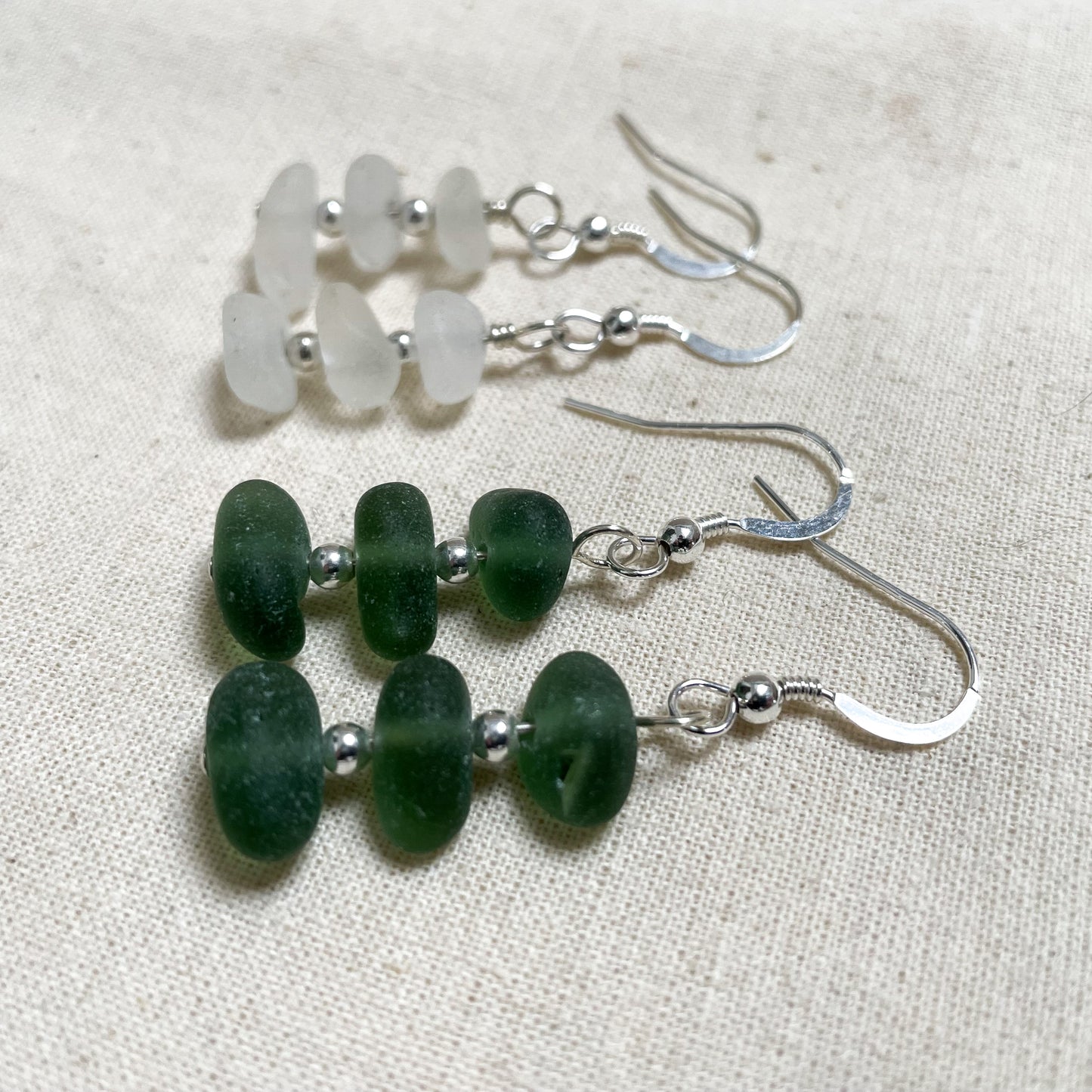 two pairs of earrings made up of three pieces of sea glass with two small silver beads inbetween. one pair is made with green sea glass the other white sea glass.