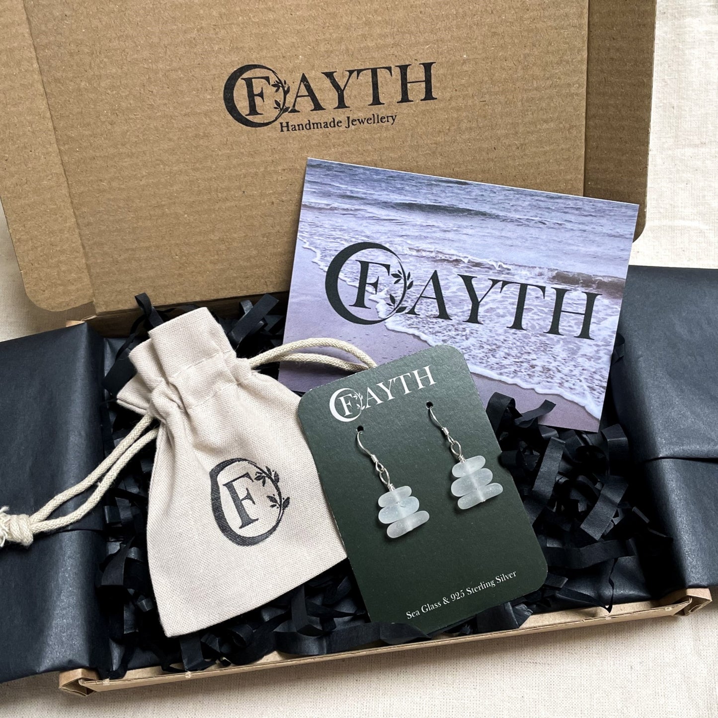 White sea glass earrings on a branded dark green display card inside a brown cardboard box with black tissue paper, a branded beige cotton tie gift bag and a business card.