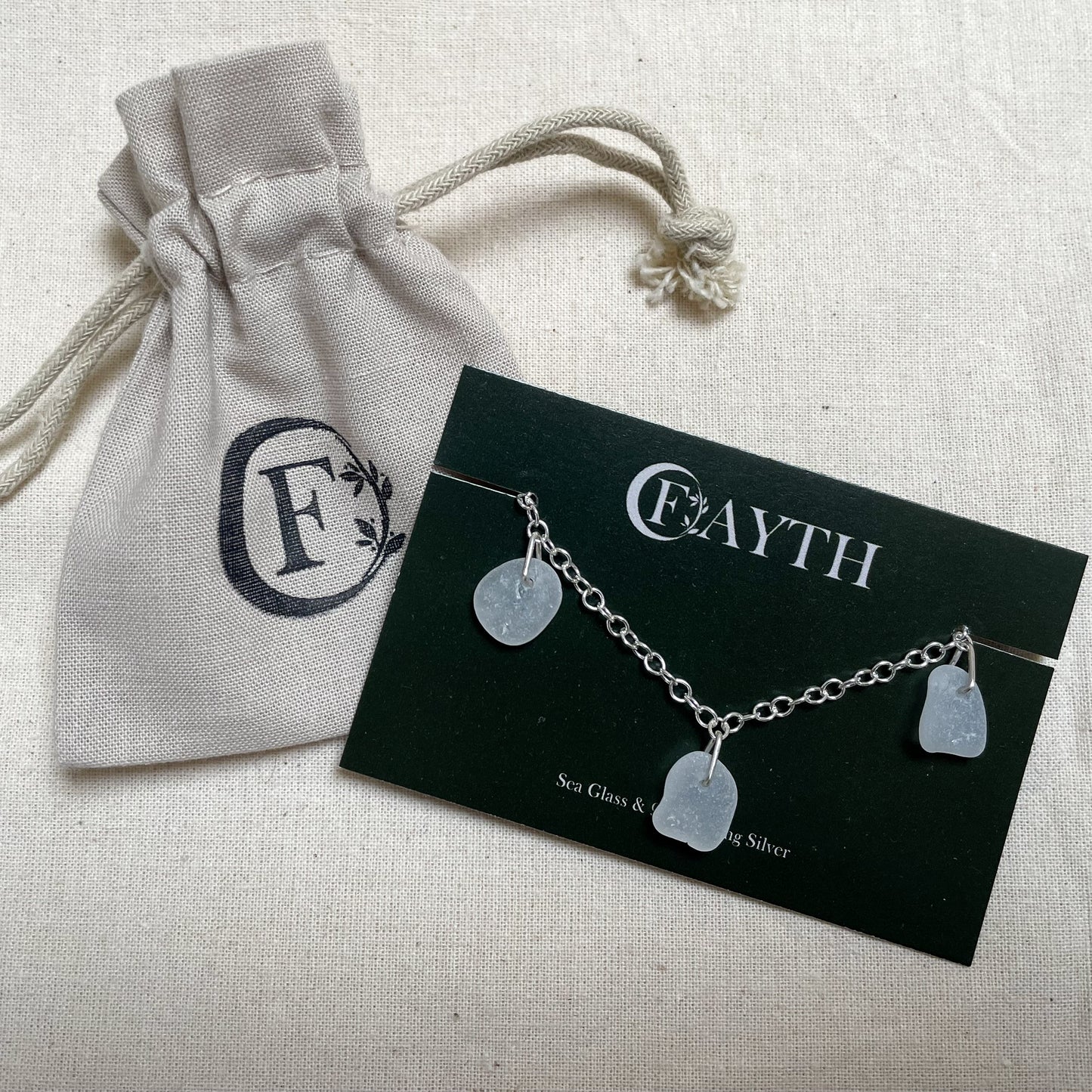 Sterling silver cable chain bracelet with three pieces of white sea glass on a dark green branded display card next to a branded beige cotton tie gift bag.