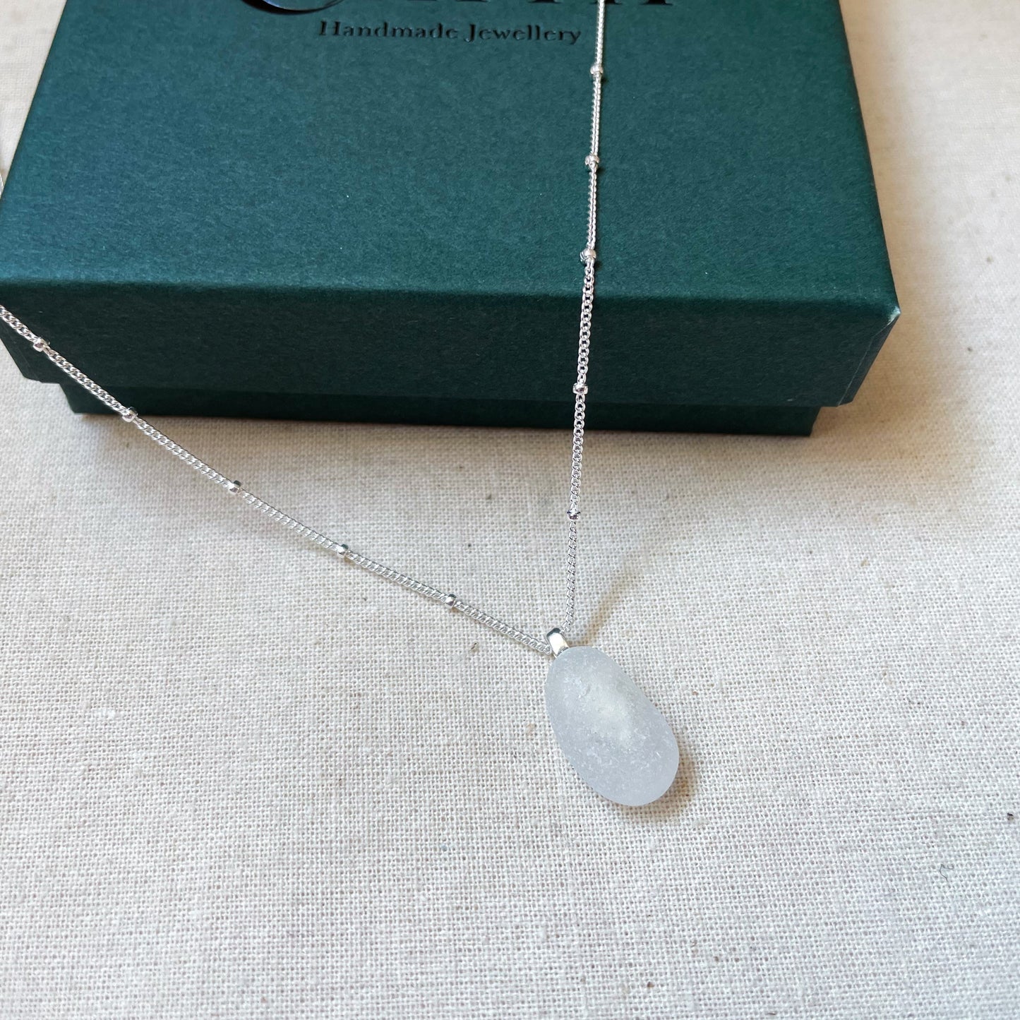 White sea glass necklace on a silver satellite chain hanging over dark green gift box