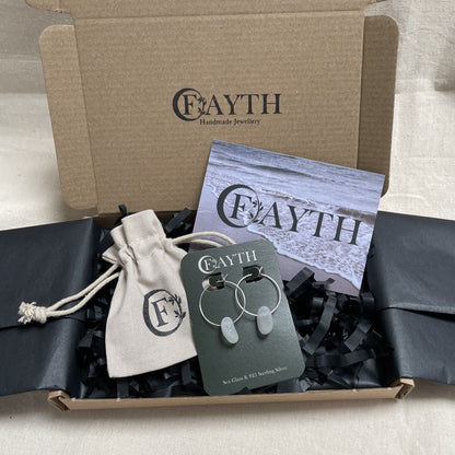 Pair of white sea glass and silver hoops, on a branded dark green display card are inside a brown cardboard box with black tissue paper, a branded beige cotton tie gift bag and a business card.