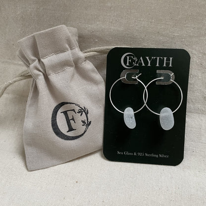 White sea glass hoops are on a dark green branded display card next to a branded light beige cotton tie gift bag