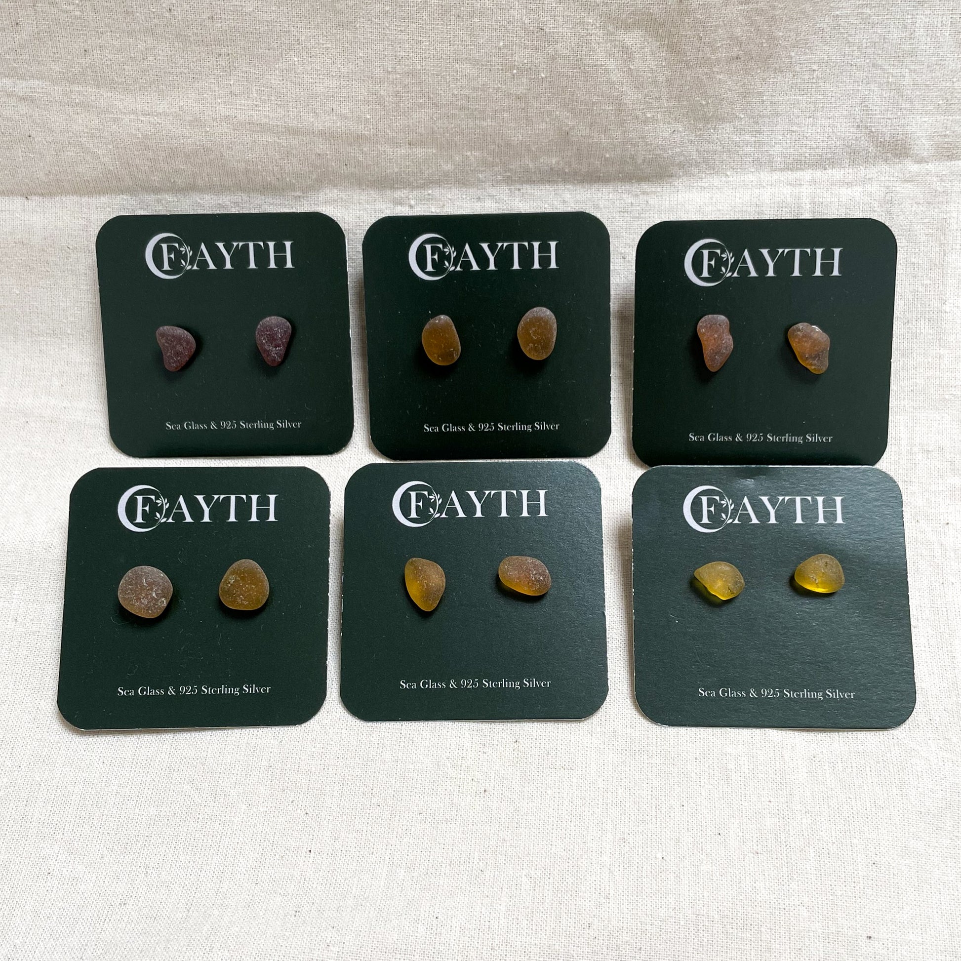 six pairs of brown/orange/yellow sea glass studs on branded dark green square display cards, in two rows of three.