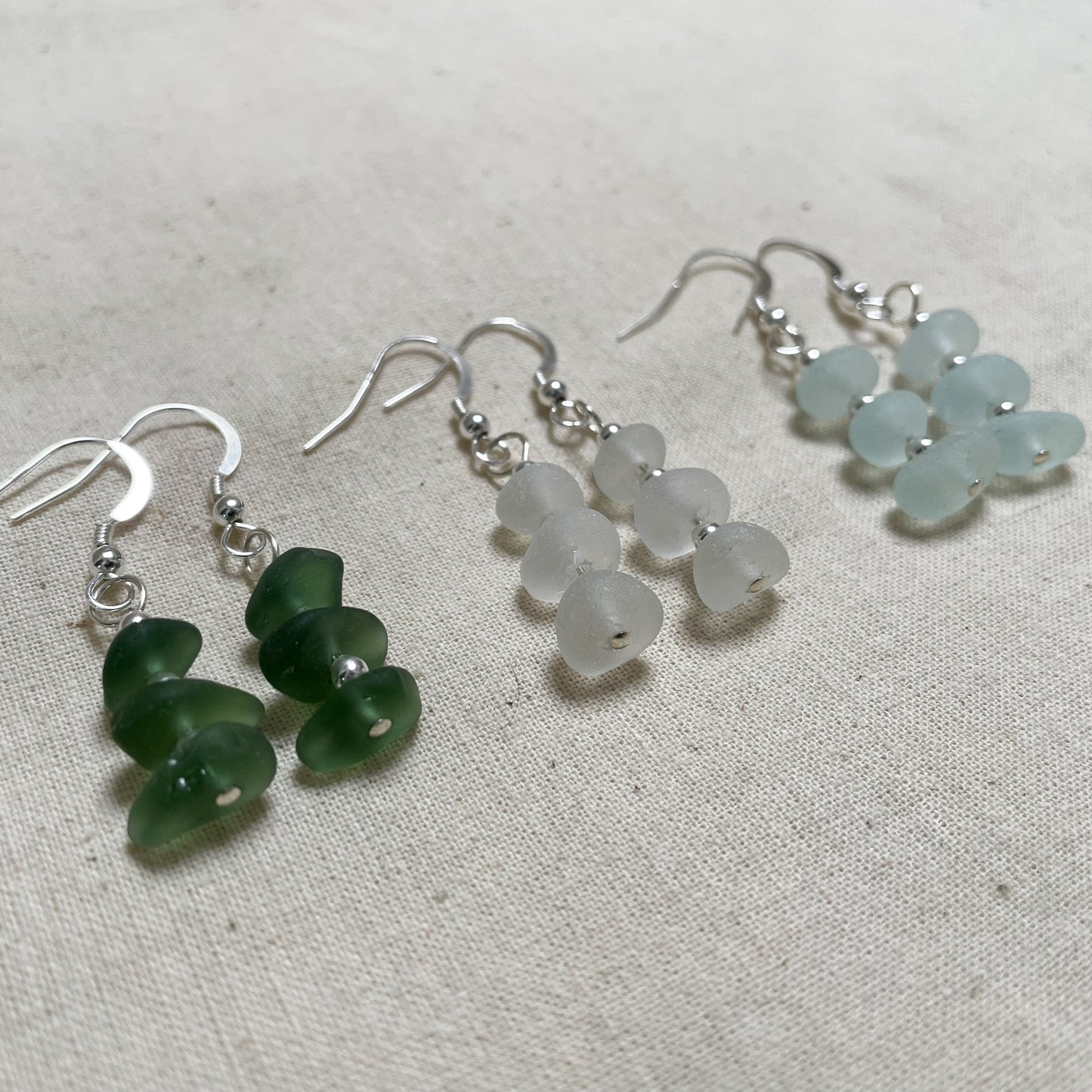 three pairs of earring in green (left), white (middle) and light blue (right). earrings are made up of three pieces of sea glass with three small silver beads in between connected to a silver ear wire with the same silver bead on