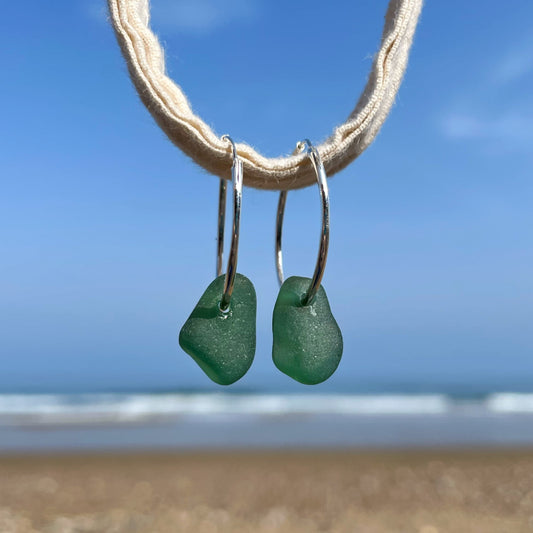 Silver sleeper hoops with green sea glass are hanging on a piece of string in front of a blurred background of the sea.