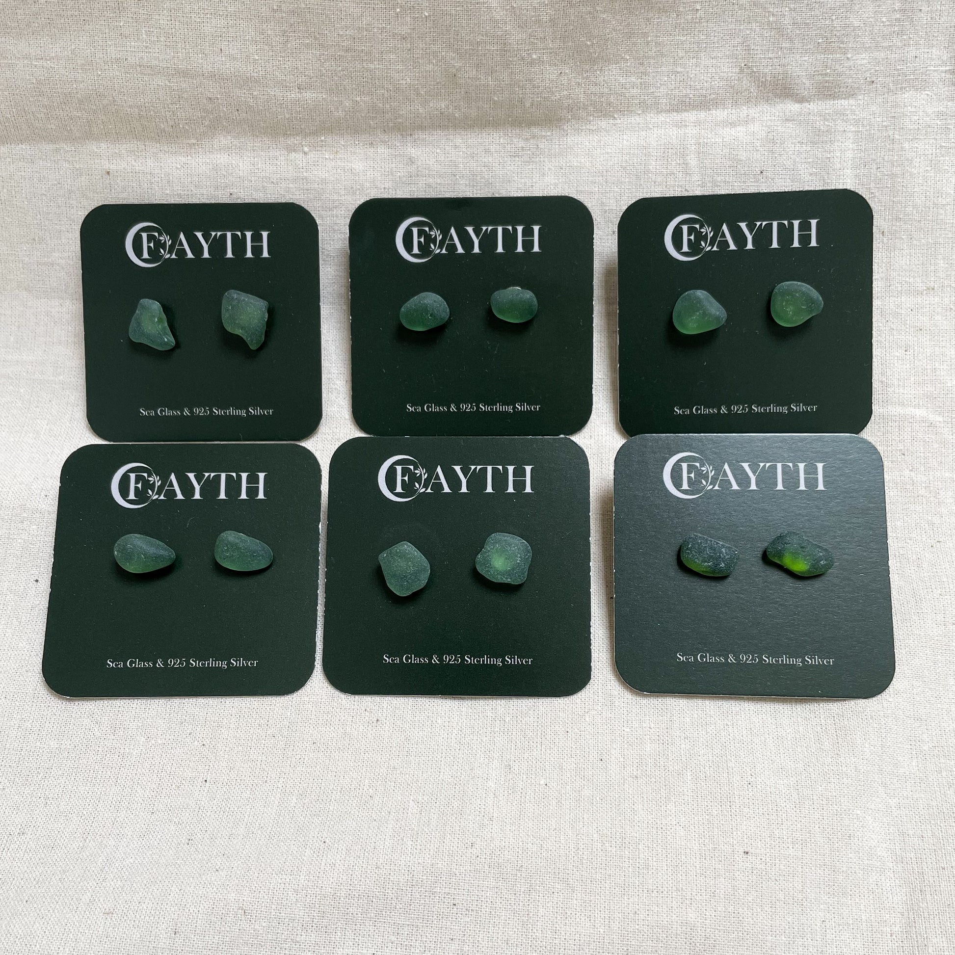 six pairs of green sea glass studs on branded dark green square display cards, in two rows of three.