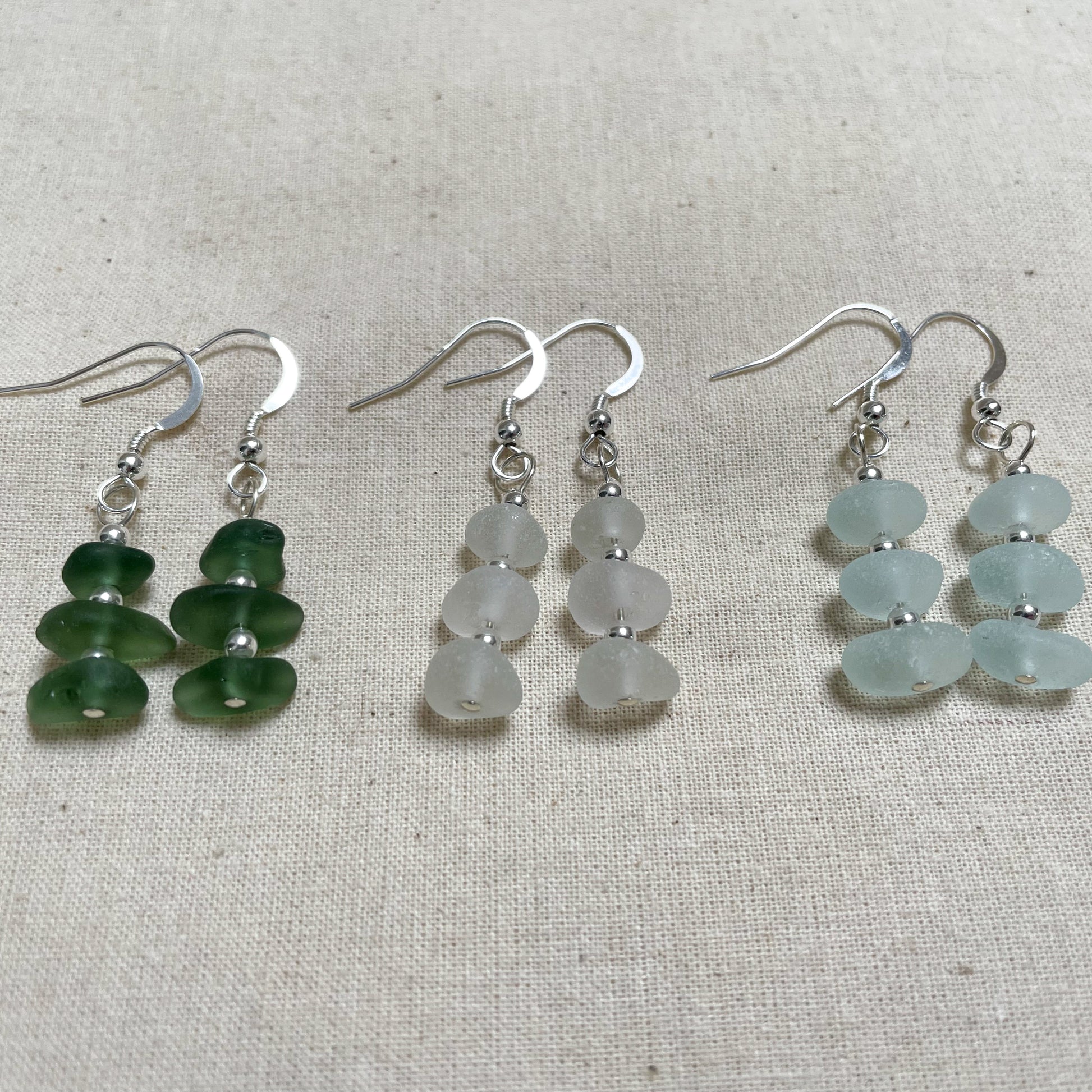 three pairs of earring in green (left), white (middle) and light blue (right). earrings are made up of three pieces of sea glass with three small silver beads in between connected to a silver ear wire with the same silver bead on