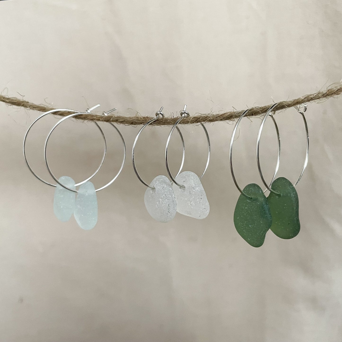 Three pairs of silver hoops with sea glass hanging from a piece of string. Two larger hoops and one smaller pair of hoops in the middle