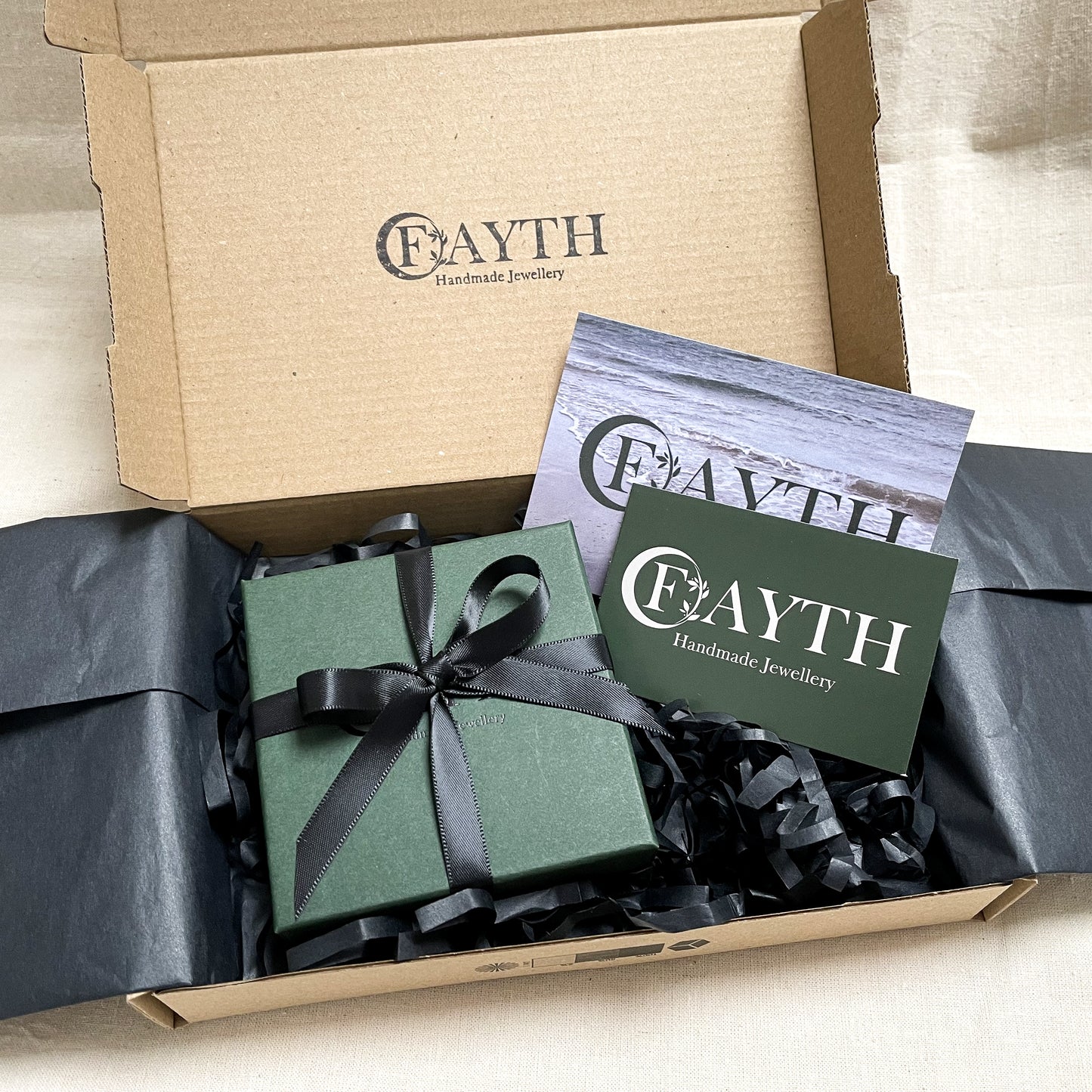 Dark green gift box with black ribbon inside a brown cardboard box with black tissue paper and business cards.