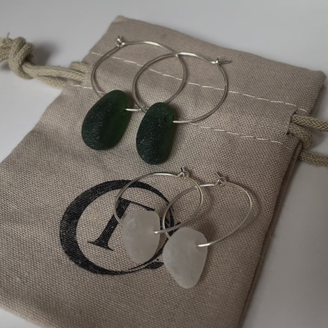 Two pairs of silver sea glass hoop earrings on a beige cotton tie gift bag. Closest pair is made with medium pieces of white sea glass and the other pair dark green sea glass.