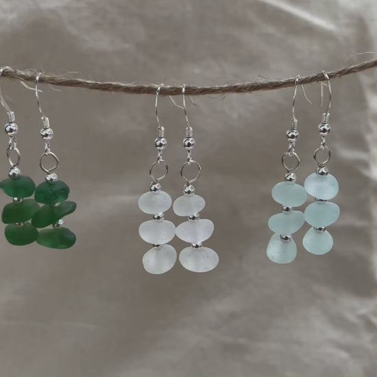 three pairs of sea glass dangle earrings hanging from a piece of string in green (left), white (middle) and  light blue (right)