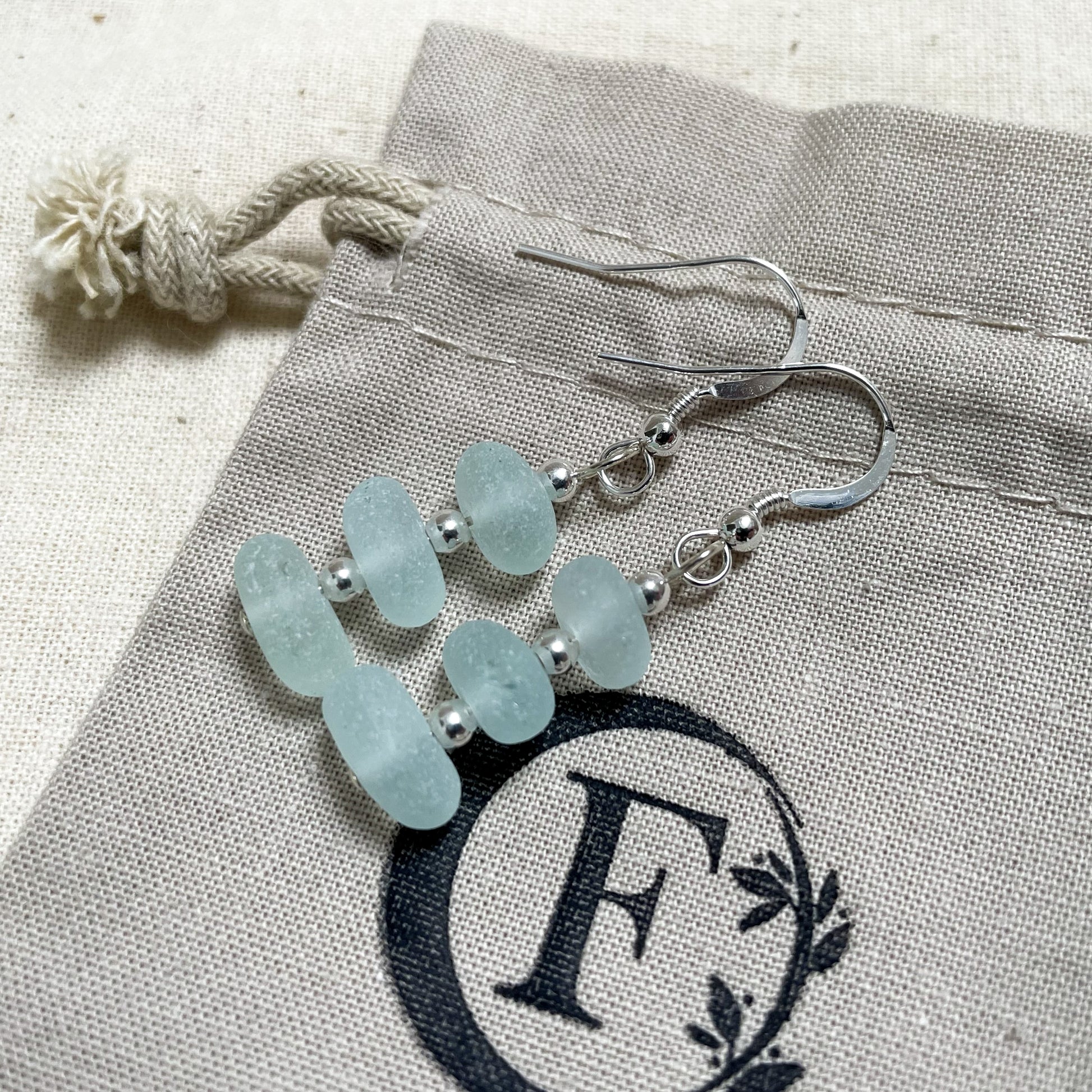 light blue seaglass and silver bead earrings laid on top of a beige cotton tie gift bag