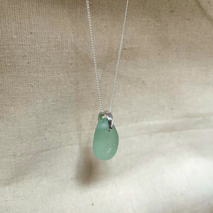 Blue sea glass necklace on fine sterling silver chain hanging against beige background.