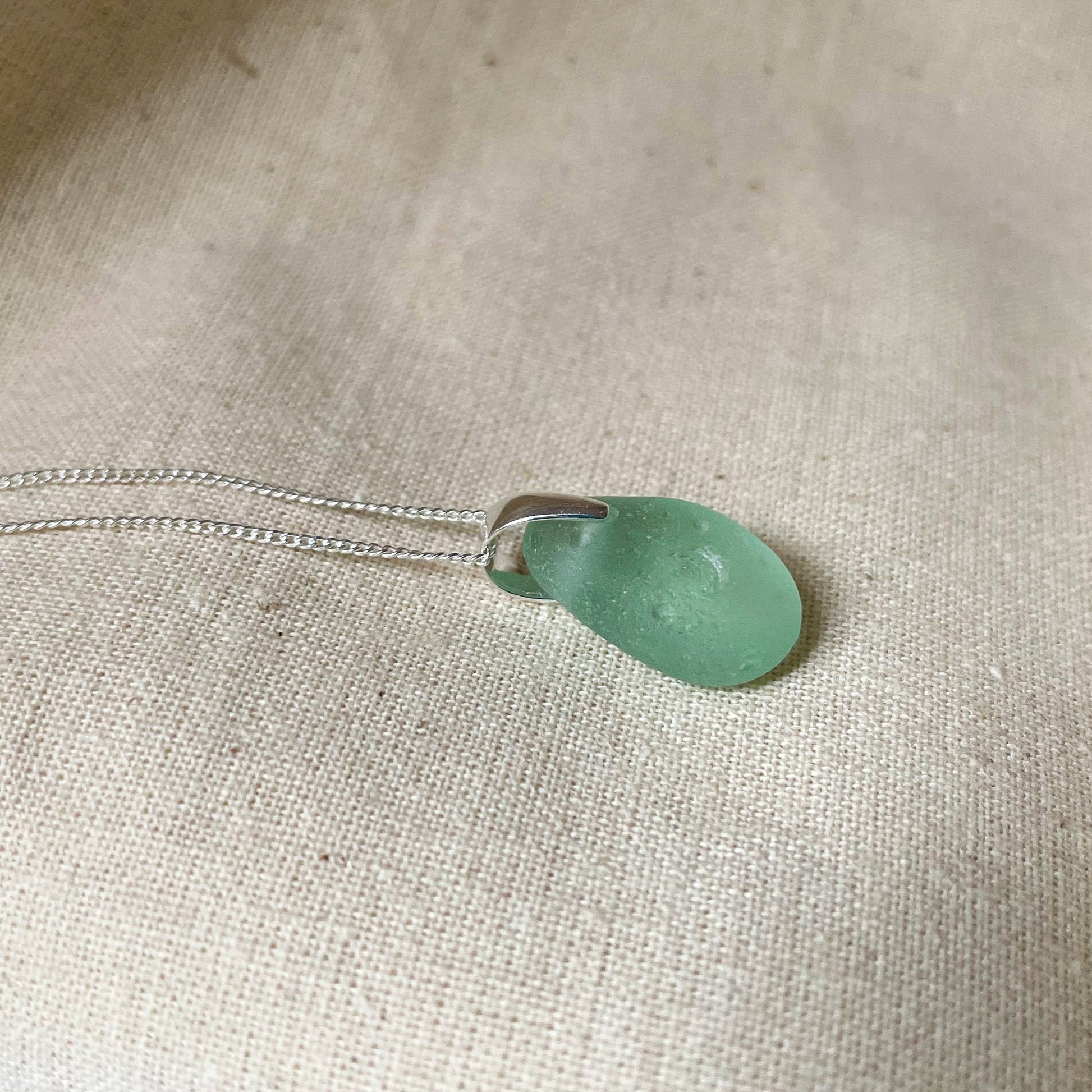 Blue sea glass pendant on fine sterling silver chain against beige background.