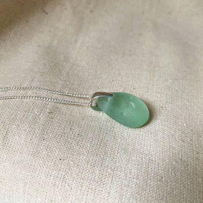 Blue sea glass pendant on fine sterling silver chain against beige background.