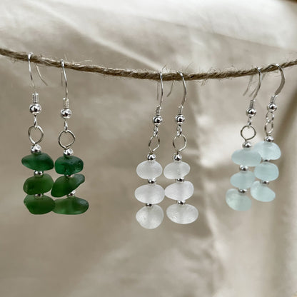 three pairs of earrings hanging from a piece of string, in green, white and light blue. earrings are made up of three pieces of sea glass with three small silver beads inbetween connected to a silver ear wire with the same silver bead on.