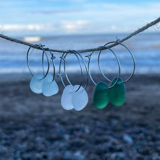 Three pairs of silver hoops with sea glass are hanging from a piece of string in front of a blurred out background of the sea.