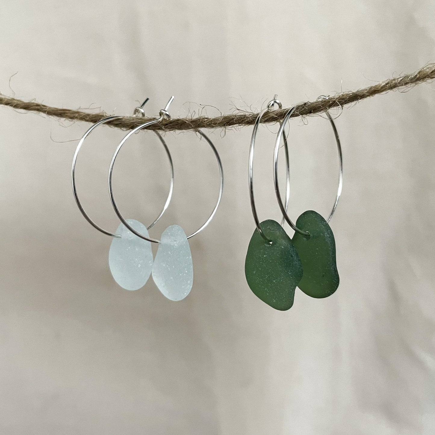 two pairs of silver hoop earrings with sea glass hanging from a piece of string. light blue sea glass hoops (left), Green sea glass hoops (right)