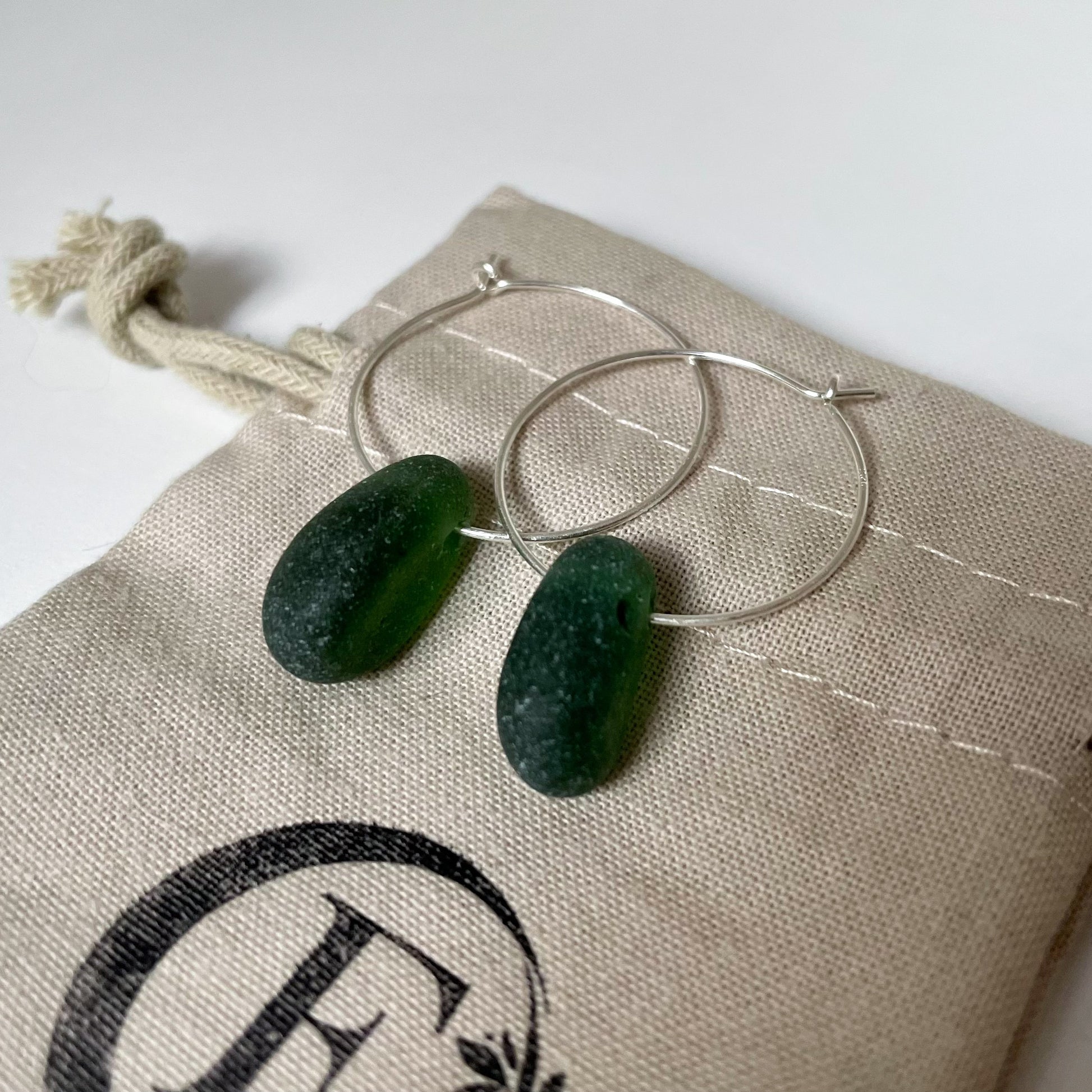 a pair of silver hoop earrings with green sea glass are laid on a beige cotton tie bag