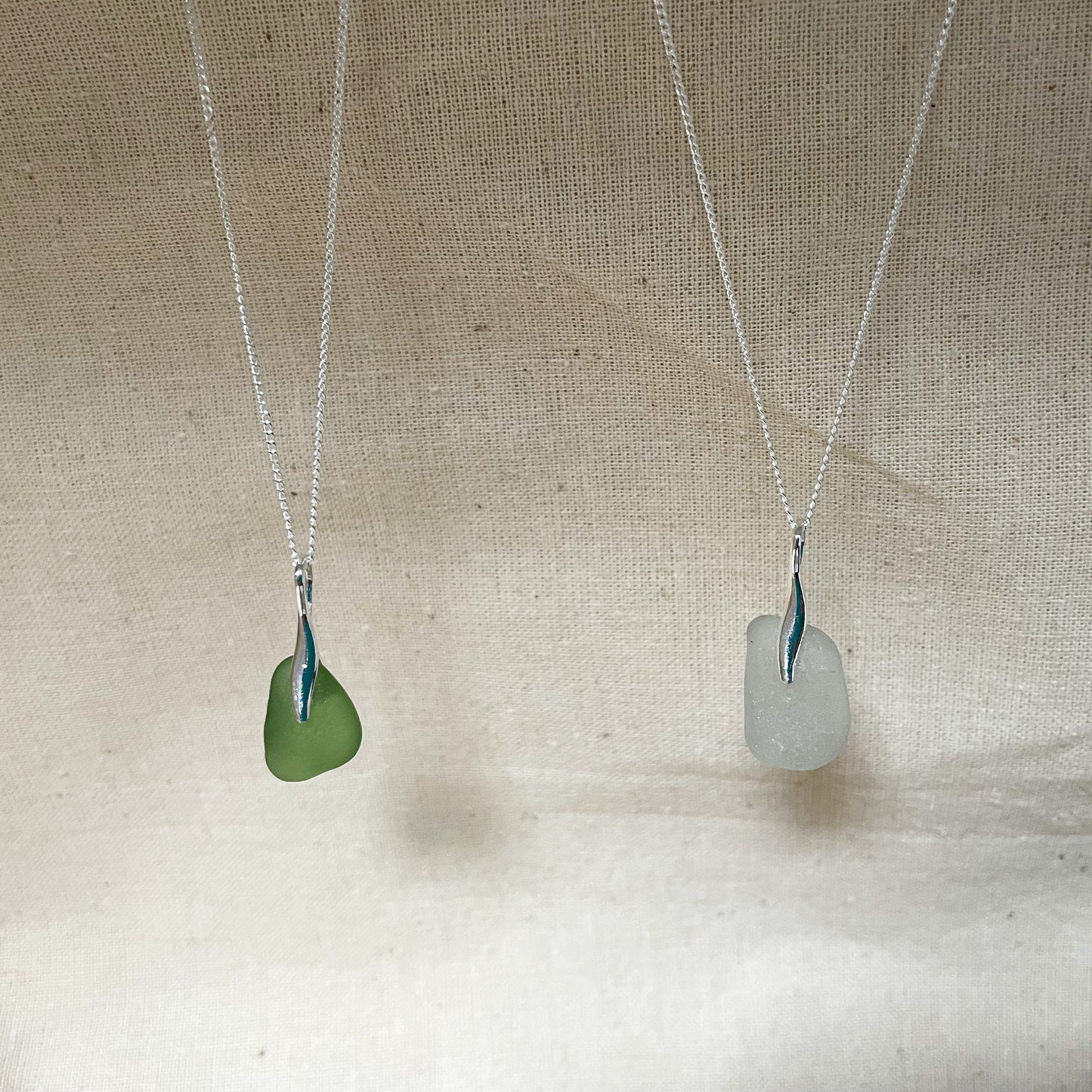 Two sea glass necklaces hanging against a beige background, green sea glass pendant on fine silver chain (left) and white sea glass pendant on fine silver chain (right)