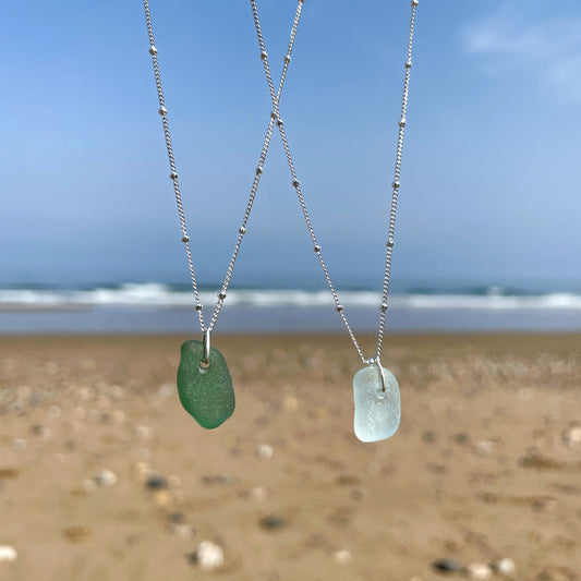 Two seaglass and sterling silver necklaces hanging in front of a blurred background of the sea. Green sea glass necklace (left) Light blue sea glass necklace (right). both necklaces are on silver satellite chains. 