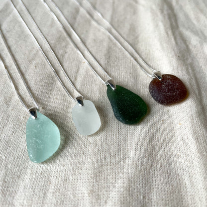 Four sea glass necklaces on fine sterling silver chains agaisnt a beige background. Light blue sea glass necklace (left), white sea glass necklace (middle), green sea glass necklace (middle) and brown sea glass necklace (right)