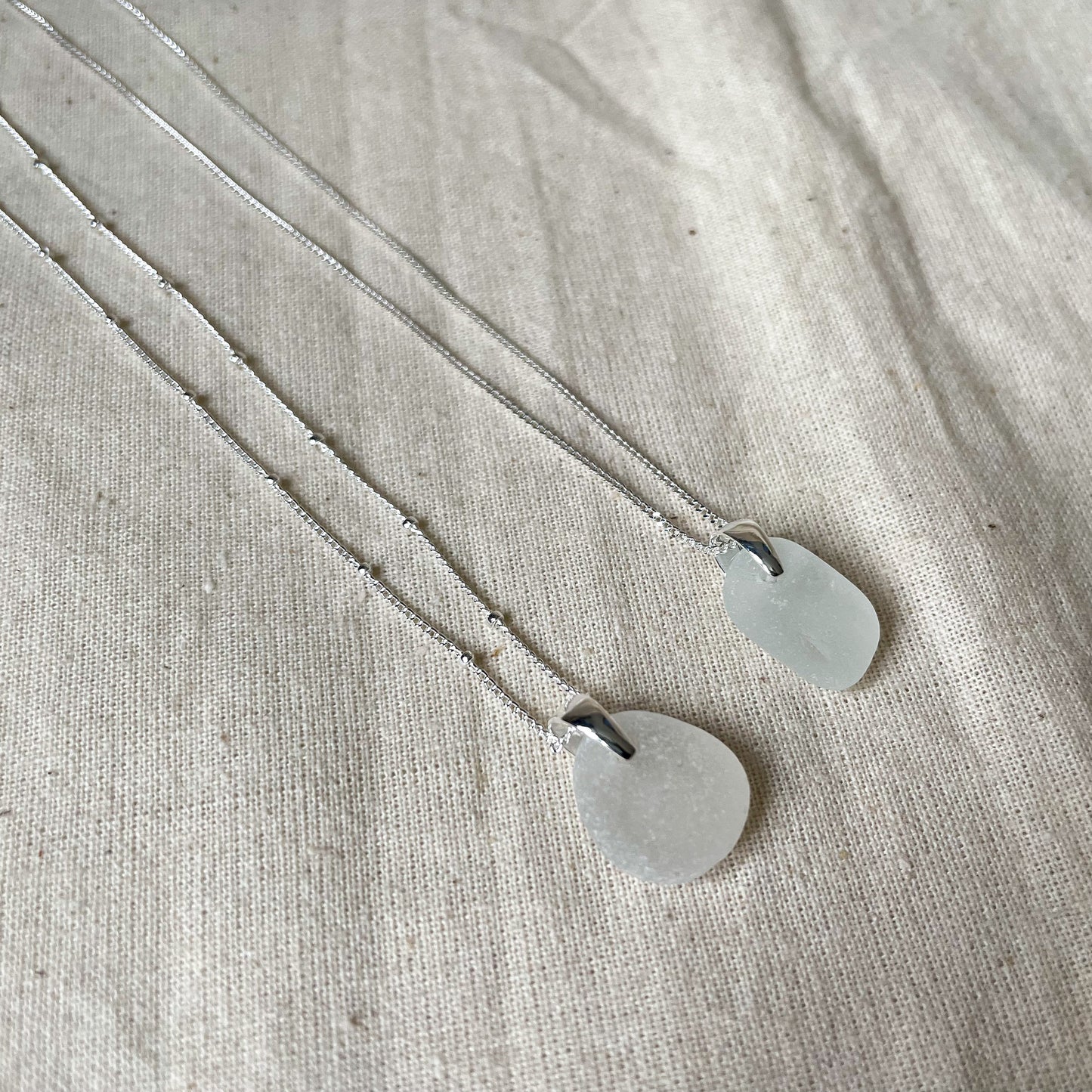 Two white sea glass necklaces on a beige background, white sea glass pendant on satellite chain (left) and white sea glass pendant on fine silver chain (right)