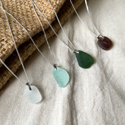 Four sea glass necklaces on fine sterling silver chains agaisnt a beige background. White sea glass necklace (left), light blue sea glass necklace (middle), green sea glass necklace (middle) and brown sea glass necklace (right)