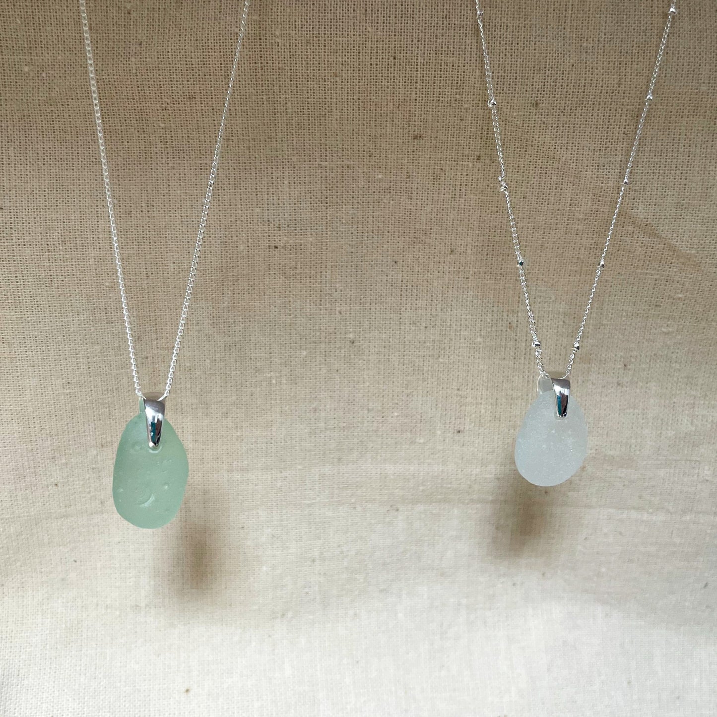 Two sea glass necklaces hanging against a beige background. Blue sea glass necklace on fine silver chain (left) and white sea glass necklace on silver satellite chain (right)