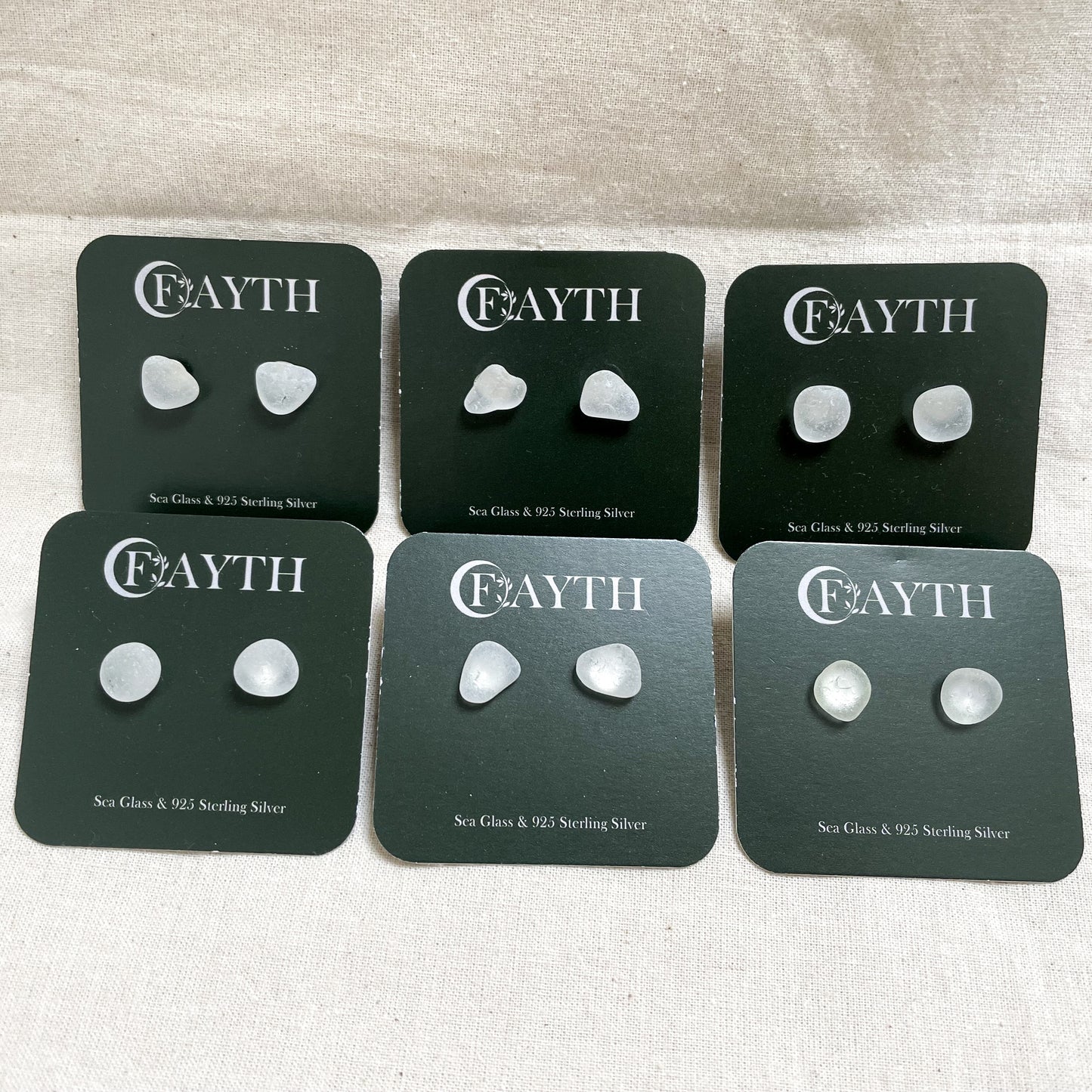 six pairs of white sea glass studs on branded dark green square display cards, in two rows of three.