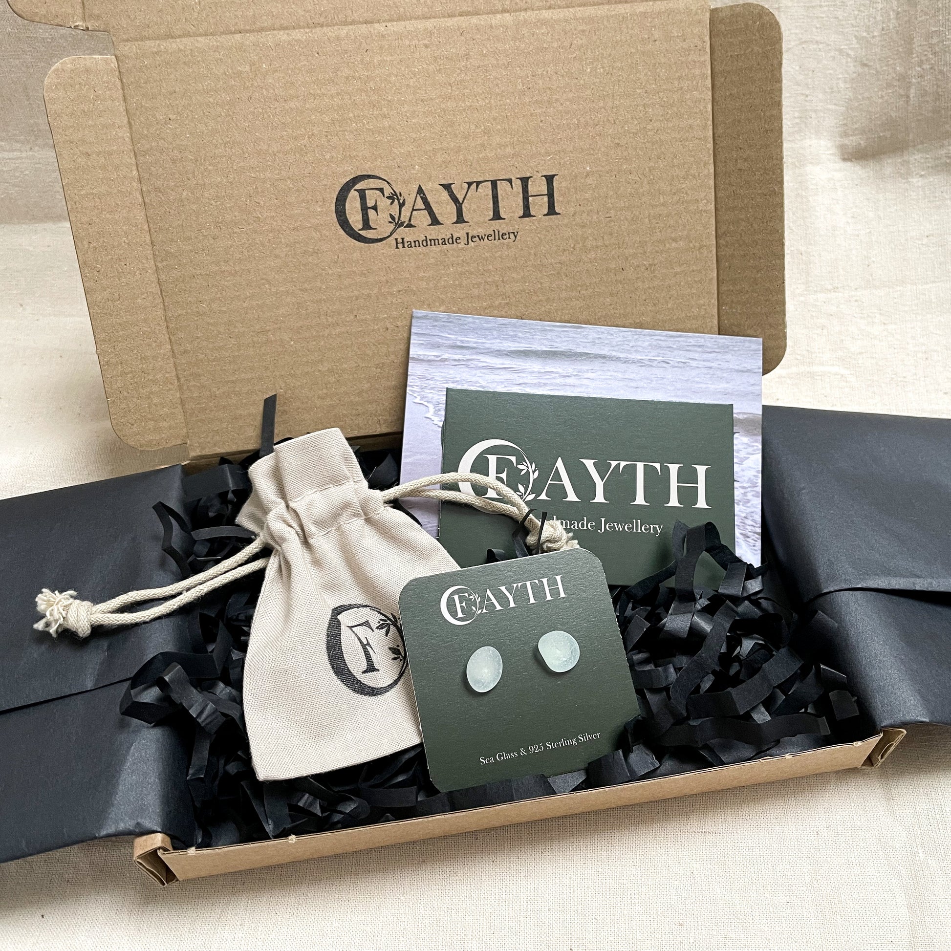 Pale blue sea glass stud earrings, on a branded dark green square display card are inside a brown cardboard box with black tissue paper, a branded beige cotton tie gift bag and a business card.